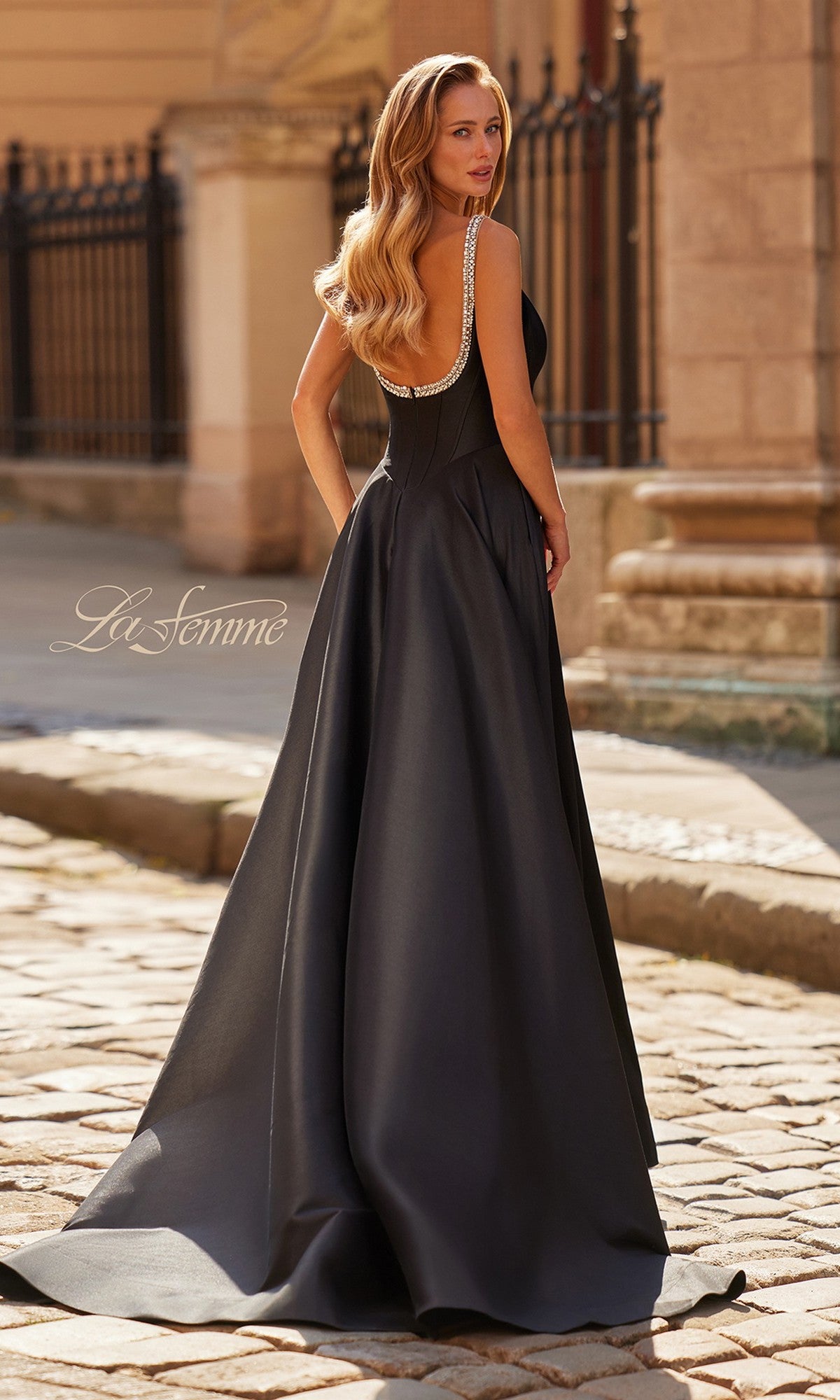 A woman with long wavy hair stands outdoors in the Long Prom Dress: La Femme 33106, a black floor-length gown featuring a crystal embellished neckline and a low, sparkling back.